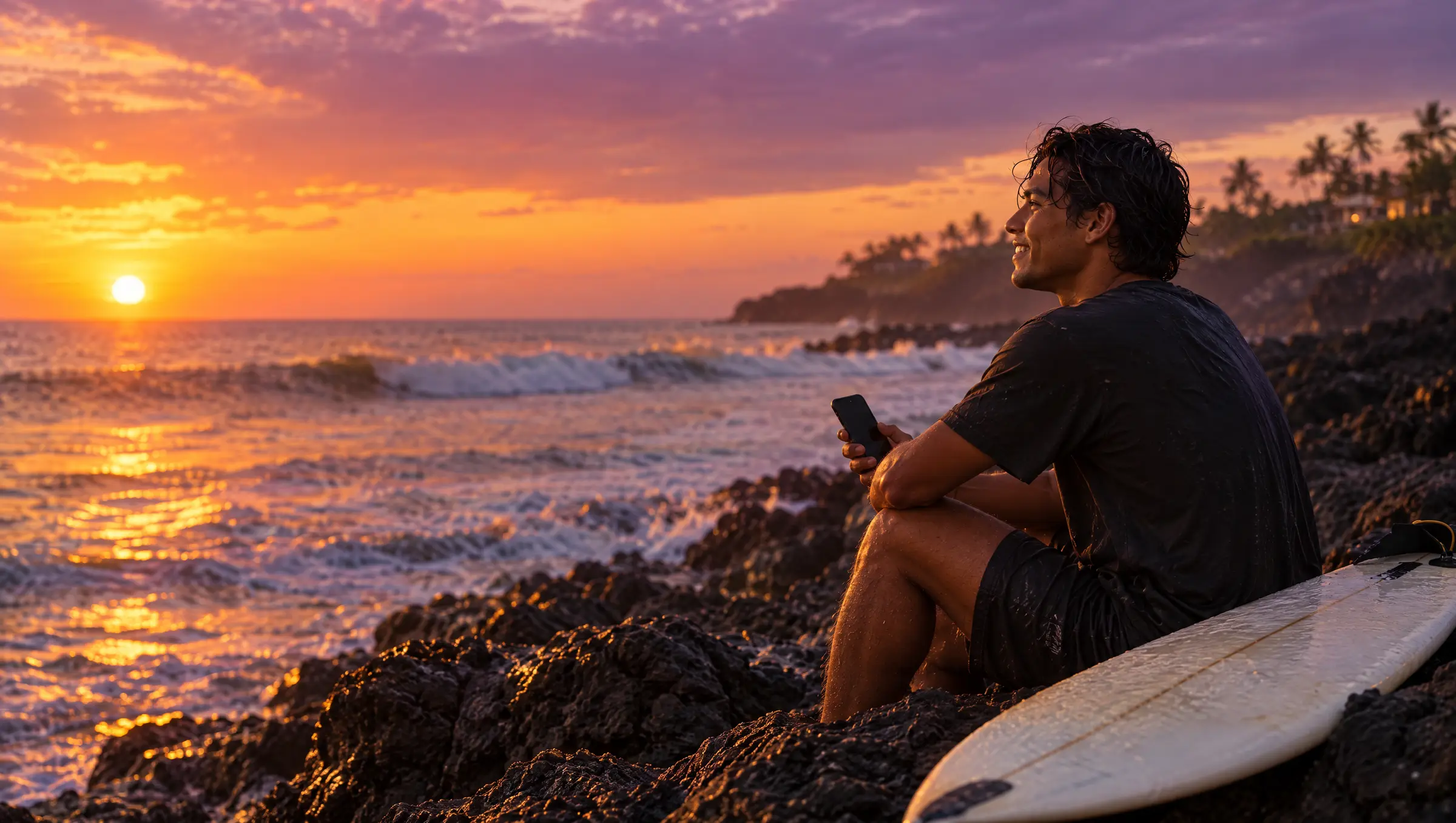 Un joven sentado en las rocas frente al mar de El Zonte, al atardecer, con su celular